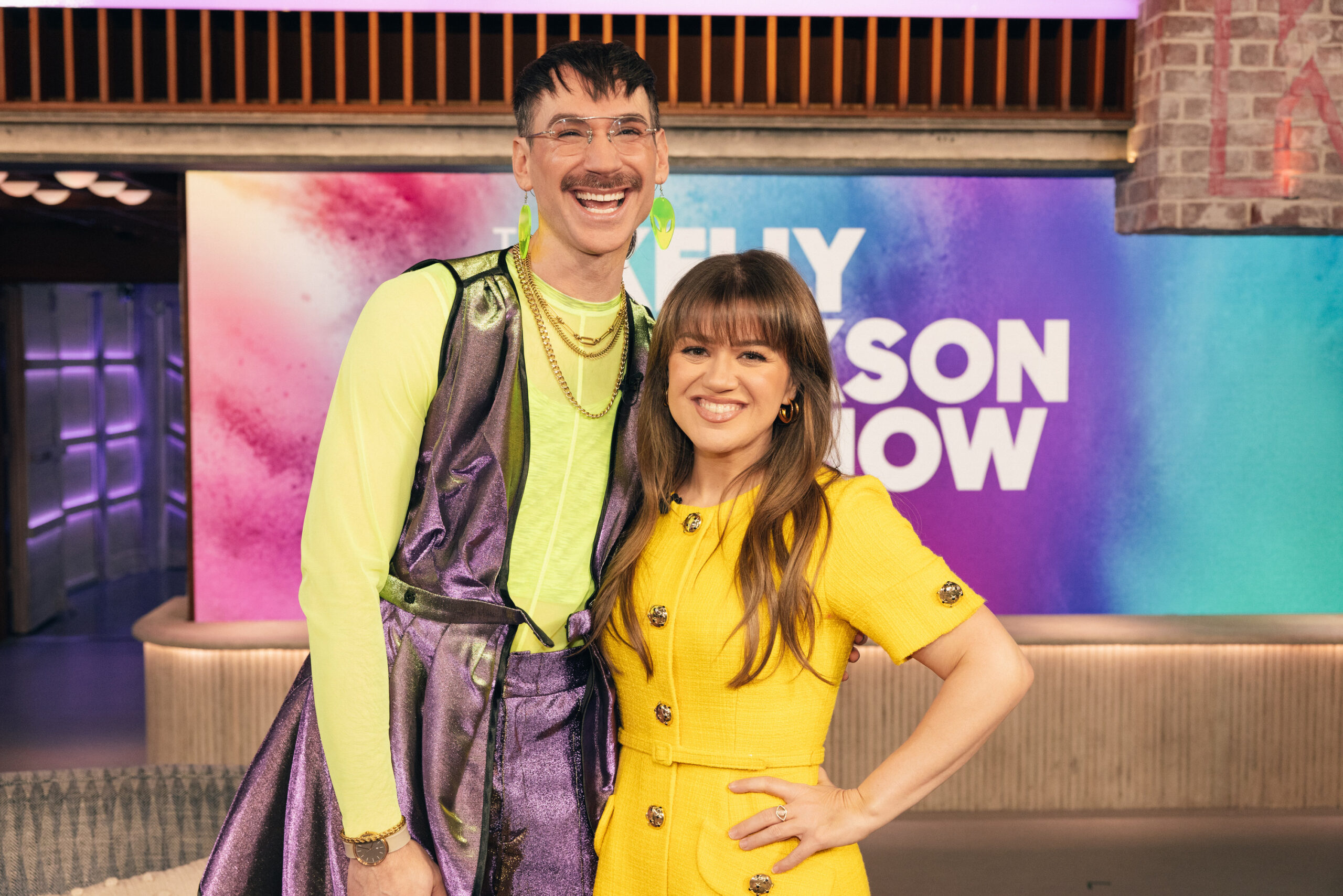 Kelly Clarkson and Kevin Gotkin are cheesing for the camera with big smiles on the set of The Kelly Clarkson Show. Kelly is a white brunette femme in a yellow dress with gold buttons and gold hoop earrings. Kevin is a white person with a mustache wearing neon green and shimmering purple, looking like a Professor of Nightlife Studies at Shiz University.